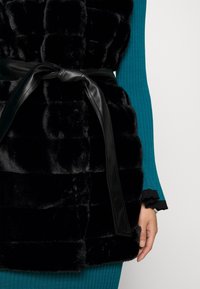 Svart faux fur-väst med ett quiltat mönster, prydd med ett slimmat svart läderbälte knutet vid midjan. Bakgrunden är vit, vilket framhäver västens textur.