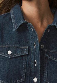 Džínová košile v tmavě modré barvě, s špičatým límcem, patentkami a náprsními kapsami. Ošoupané detaily dodávají texturu. Stříbrné kovové ozdoby.