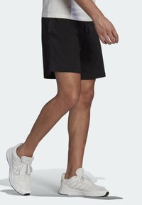 Pantalones cortos deportivos negros con una cinturilla elástica y cordón, combinados con zapatillas blancas que tienen una superficie texturizada y un logo prominente.
