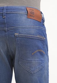Blå denimjeans med en läderlapp märkt "RAW." Funktioner inkluderar en bakficka med dekorativ sömnad och en mjuk textur.