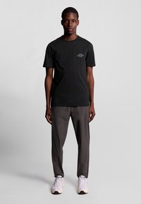 Sort kortærmet t-shirt med grafisk tryk på brystet, parret med grå bukser og lyse sneakers, stående mod en ensfarvet baggrund.