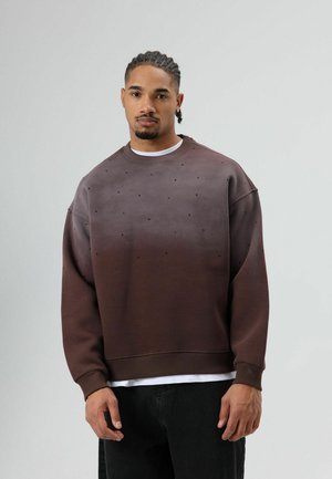 Bruine sweatshirt met een verloopkleur en een textuur met kleine zwarte stippen. Losse pasvorm en geribbelde manchetten, gedragen over een wit T-shirt.