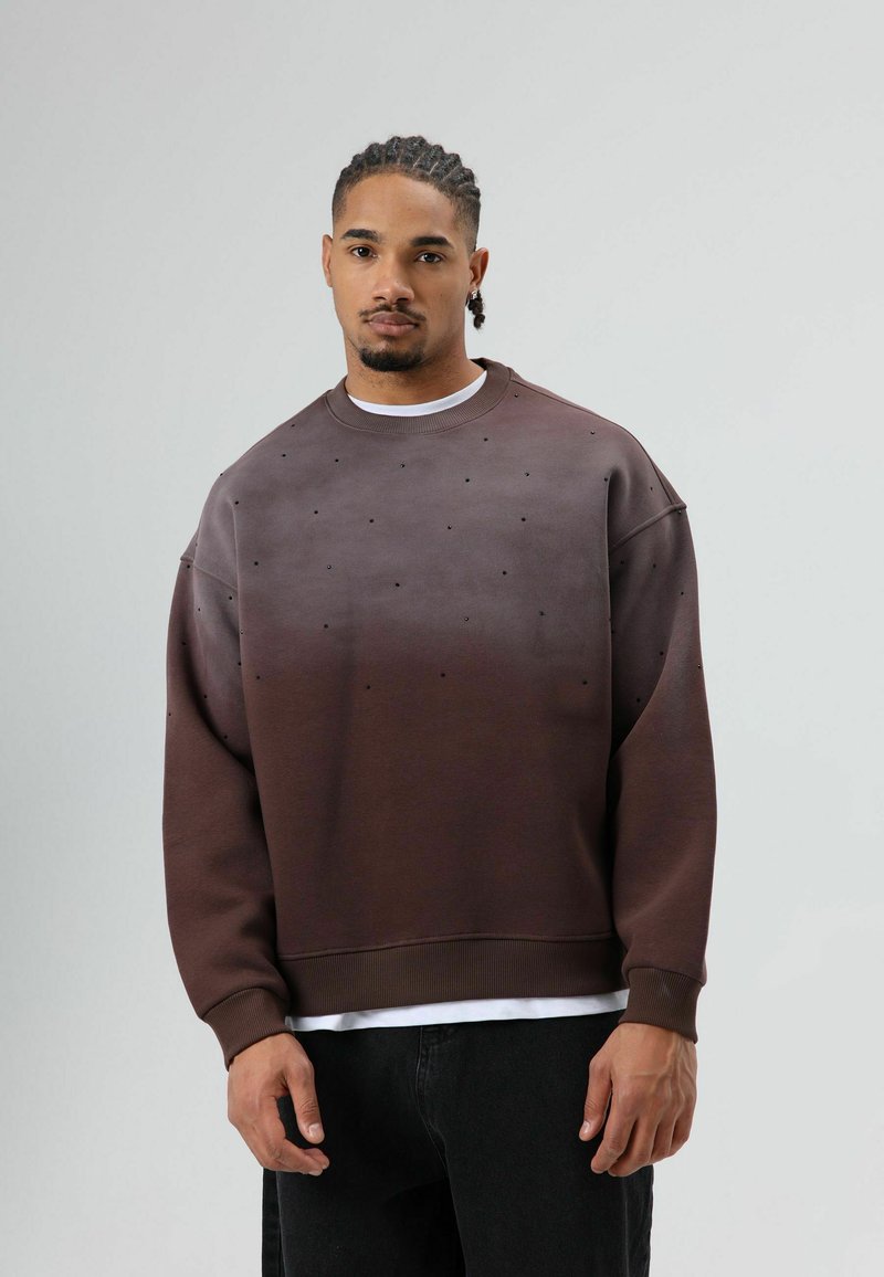 Bruine sweatshirt met een verloopkleur en een textuur met kleine zwarte stippen. Losse pasvorm en geribbelde manchetten, gedragen over een wit T-shirt.