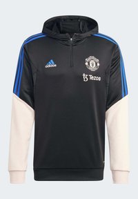 Felpa nera Adidas con strisce blu sulle spalle e maniche color crema. Emblema del Manchester United e logo Tezos sul fronte. Design a mezza zip.