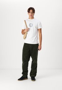 Witte katoen T-shirt met een blauw grafisch ontwerp, gecombineerd met donkergroene corduroy broek. Zwarte sneakers met een rubberen zool en een beige schoudertas.