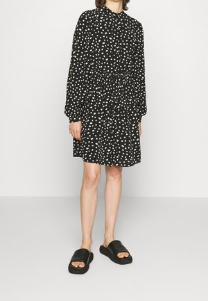 Person iført en sort, langærmet kjole med hvidt blomsterprint, markeret talje og sorte åbne slidesandaler, stående på en ensfarvet gulv.