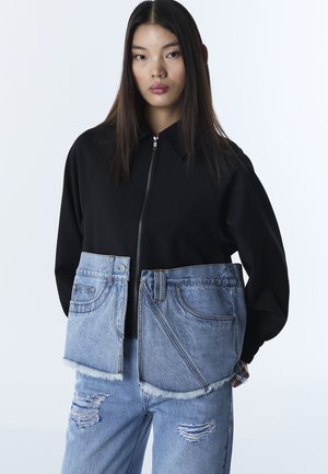 Młoda kobieta ubrana w czarną bluzę na zamek, na którą nałożona jest oversize'owa niebieska kamizelka dżinsowa oraz podarte, jasnoniebieskie jeansy, na tle jednolitego białego tła.