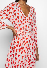 Frau, die ein langärmliges, v-ausschnitt Wickelkleid mit roten Herzmustern und einem gebundenen Gürtel in der Taille auf einem hellrosa Stoff trägt.