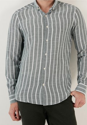 Man die een lichtgrijs-witte gestreepte blouse met lange mouwen en een zwarte broek draagt, staand tegen een effen achtergrond.