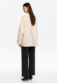 Mujer con cabello largo vestida con una camisa crema oversize, pantalones anchos negros y botines de tacón negros, de pie contra un fondo blanco.