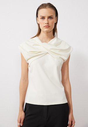 Kurze, weiße Bluse mit einem verdrehten Stoffdesign am Ausschnitt, glatter Textur und taillierter Form, kombiniert mit schwarzen Hosen.