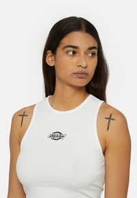 Witte mouwloze crop top van katoen met een ronde halslijn. Gemaakt met een zwart "Dickies" logo in het midden van de borst. Licht getailleerd ontwerp.