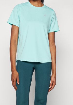 Femme portant un t-shirt de sport bleu clair à manches courtes et un legging bleu sarcelle foncé, debout devant un fond blanc uni.