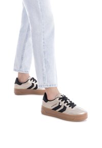 Zapatillas con una parte superior beige metálica, acentos en negro y tres rayas negras. Presentan una suela texturada en color marrón y cordones negros.