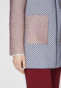 Textured jacket with a geometric blue and white pattern and a contrasting pink netted sleeve. Features a front pocket and zip closure.