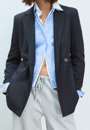 Persoon draagt een zwart blazer over een open blauwe gestreepte blouse die een bloot middel onthult en lichtgrijze joggingbroek met trekkoord.