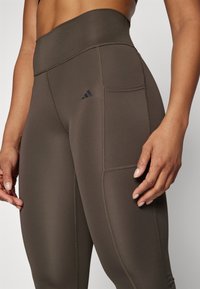 High-waisted, dark brown leggings made of stretch fabric with a smooth texture, featuring a side pocket and black logo accent.