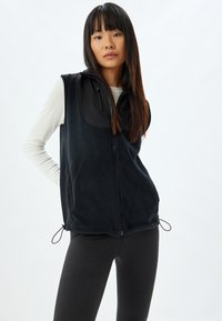Gilet in pile nero con zip frontale, presenta un pannello in tessuto nero sul petto e cordini regolabili all'orlo per una vestibilità personalizzata.