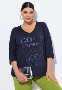 Navyblaues V-Ausschnitt-Oberteil mit drei Viertelärmeln und metallischem Text. Kombiniert mit grünen Hosen hält das Model eine silberne Clutch.