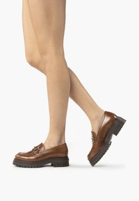 Mocassins en cuir marron avec bout arrondi, boucle décorative et semelle en caoutchouc noir épaisse. Texture lisse avec des détails de surpiqûres sur les côtés.