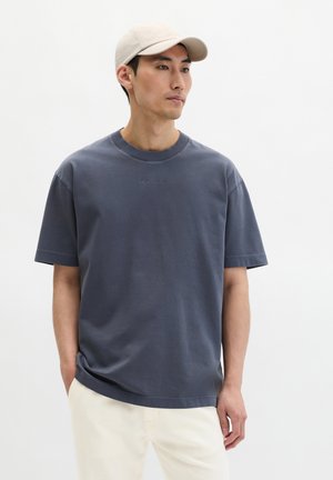 Mann trägt ein graues T-Shirt mit kurzen Ärmeln, beige Hose und eine hellbeige Baseballkappe, steht mit einer Hand in der Tasche.