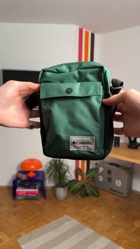 Zelená látková taška s klopou a boční kapsou, s černým spodkem a stříbrným logem Columbia na přední straně. Pevný design a textura.