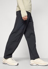 Mustat urheiluhousut, joissa on slim fit -malli, kevyestä kankaasta valmistetut, näkyvillä saumoilla ja taskuilla, yhdistettynä vaaleansävyisiin juoksukenkiin.