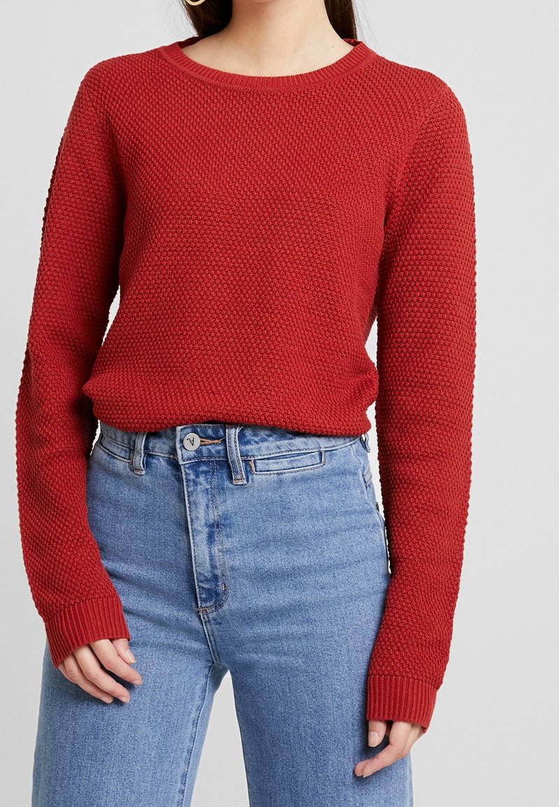 Pull texturé rouge avec un col rond et des manches longues, associé à un jean taille haute bleu clair. Poignets côtelés visibles.