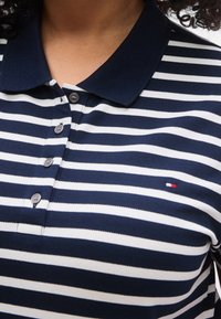 Πουκάμισο πόλο με ρίγες σε ναυτικό μπλε και λευκό, με γιακά, τρία κουμπιά και ένα μικρό λογότυπο σε κόκκινο, λευκό και μπλε στο στήθος.
