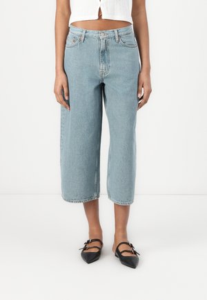 Femme portant un jean large et court bleu clair associé à des ballerines noires pointues à double bride sur fond blanc.
