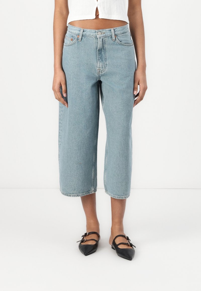 Femme portant un jean large et court bleu clair associé à des ballerines noires pointues à double bride sur fond blanc.