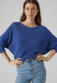 Modra pletena jopica ohlapnega kroja, širokega izreza in rebraste teksture, v kombinaciji z visokim pasom svetlikavimi jeans hlačami.