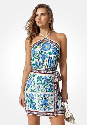 Žena nosi šaty s halterovým výstrihom s modrými kvetinovými vzormi a geometrickými okrajmi, v pravej ruke drží malú tkanú kabelku.