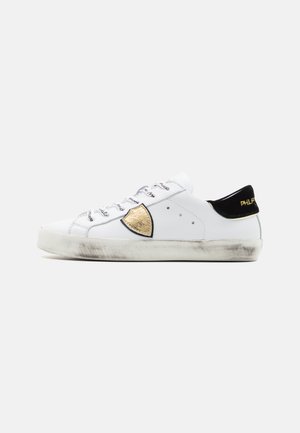 Sneaker bianca con accenti neri e dorati, pannello dorato testurizzato, suola grigia chiara e lacci con motivo. Leggero usura sulla suola.