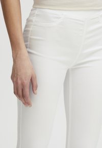 Pantalones de denim elástico blanco con una textura suave, que presentan bolsillos frontales y un diseño ajustado. Mano reposando en la zona del muslo.