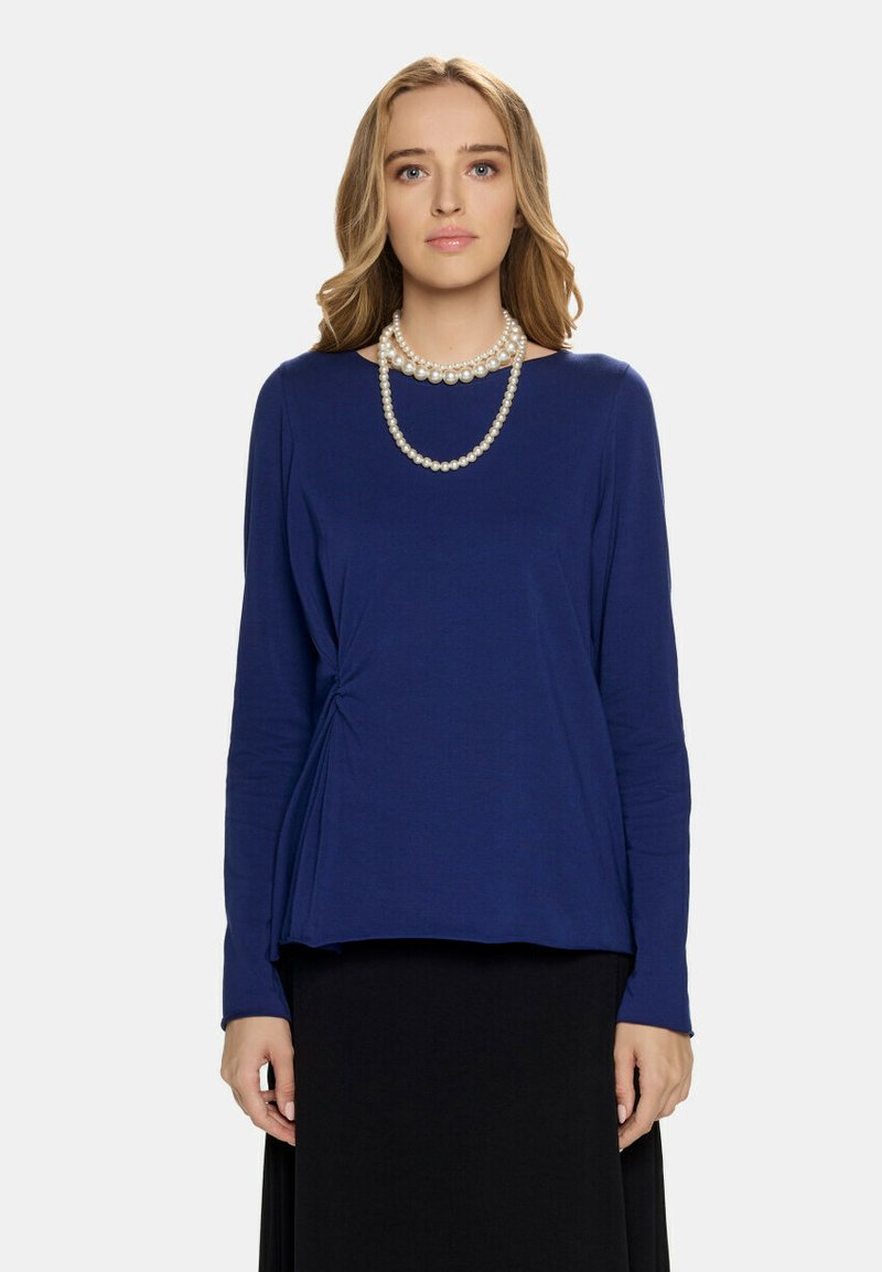 Granatowa bluzka z długim rękawem z skręconym detalem na boku, o miękkiej fakturze i luźnym kroju, zestawiona z czarną spódnicą i warstwowym naszyjnikiem z pereł.