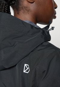 Black waterproof jacket with a textured surface, featuring a logo on the shoulder, adjustable drawstring hood, and streamlined design details.