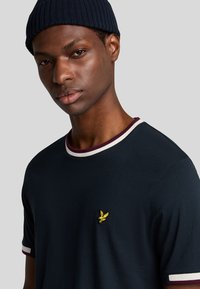 Maglietta blu navy con collo tondo, rifiniture a coste bordeaux e bianche in contrasto, e un piccolo logo ricamato giallo sul petto.