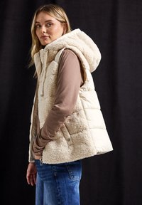 Gilet en polaire beige avec capuche, fermeture éclair et poches latérales. L'intérieur est lisse, tandis que l'extérieur présente une surface douce et texturée.