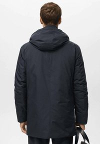 Cappotto imbottito blu navy con cappuccio, caratterizzato da una vestibilità dritta, texture liscia e maniche lunghe. Indossato sopra pantaloni scuri, mentre tiene una tracolla nera.