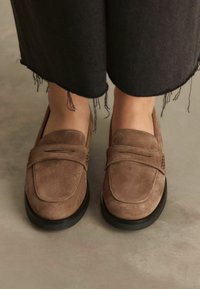 Mocassini in suede marrone con punta arrotondata, caratterizzati da una banda cucita sulla parte anteriore e una suola in gomma nera testurizzata.