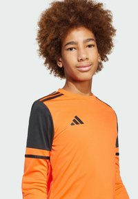 Oranje sportshirt met lange mouwen en zwarte accenten en strepen op de schouders. Bevat het zwarte Adidas logo op de borst. Gladde stoftextuur.