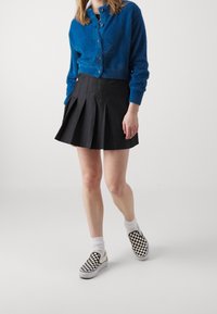 Niebieski, puszysty kardigan, czarna plisowana spódnica i czarno-białe buty wsuwane w kratkę noszone z białymi skarpetami. Proste, teksturowane materiały.