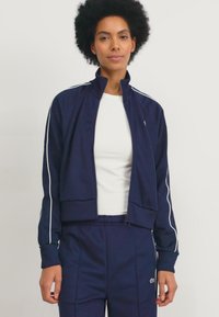 Giacca zip-up blu navy con strisce bianche, polsini a coste e design cropped. Abbinata a un top bianco e pantaloni blu navy coordinati.