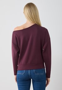 Sudadera burdeos sin hombros, con mangas largas y dobladillo acanalado, combinada con jeans azules. Tejido suave con un ajuste relajado.