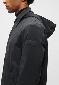 Fekete vízálló dzseki kapucnival, patentos gallérral, oldalzsebekkel és egy texturált logófolttal a bal ujján.