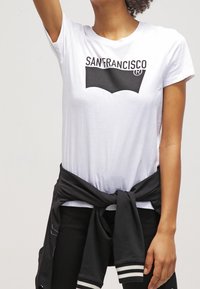 Persona vistiendo una camiseta blanca con la inscripción "SAN FRANCISCO" y una chaqueta negra atada a la cintura, de pie con un brazo levantado.
