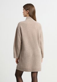 Vestido de punto beige con un cuello alto, ajuste holgado y puños acanalados. Textura suave, longitud hasta mitad del muslo y diseño minimalista.