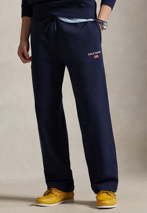 Tmavomodré tepláky s elastickým pásom so šnúrkou, s nápisom "POLO SPORT" a detailom americkej vlajky. Nosené so žltými topánkami.