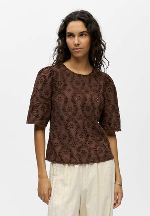 Kiharatummatukkainen nainen pukeutunut ruskeaan tekstuuriin, lyhythihaiseen puseroon, jossa on kukkakuosia, ja vaalean beigeihin housuihin seisoo valkoista taustaa vasten.